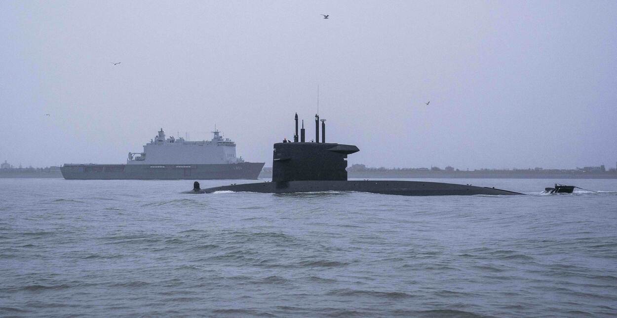 Onderzeeboot half boven water met op de achtergrond een marineschip.