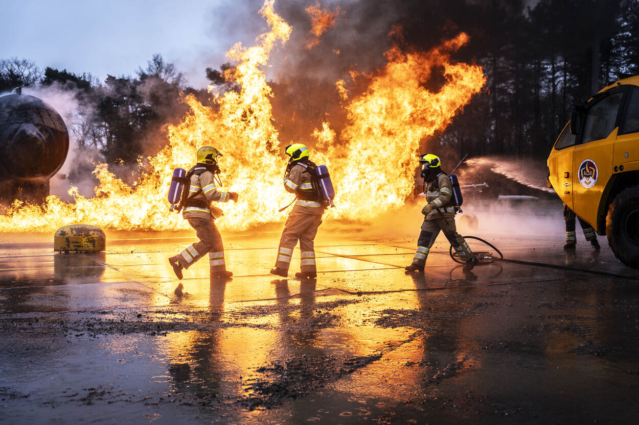Enorme felgele en oranje vlammen schieten uit de mock-up van een verkeersvliegtuig met op de achtergrond gitzwarte rookwolken.