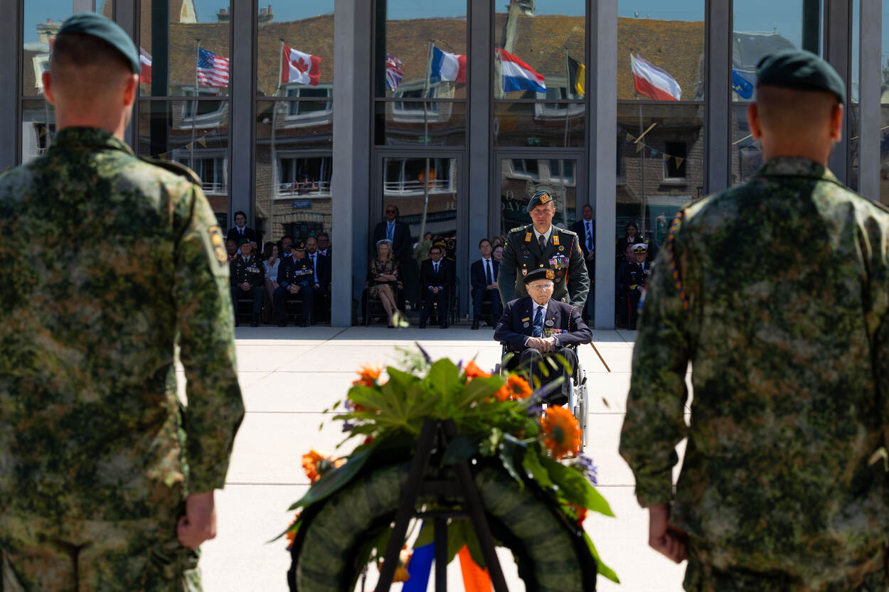 Tweede Wereldoorlogveteraan Max Wolff wordt in een rolstoel richting een bloemenkrans gereden door een luitenant-kolonel.
