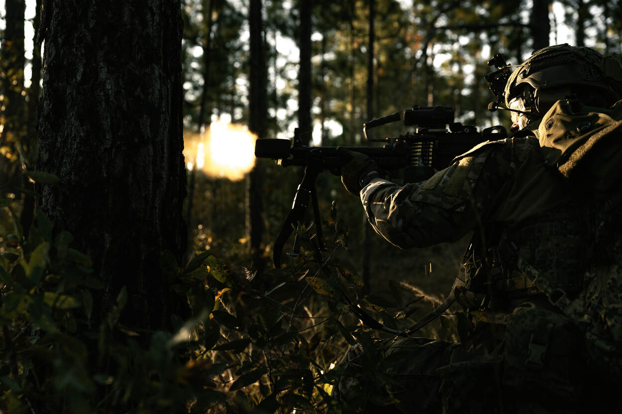 Een militair zit in en bos en vuurt met zijn wapen. Door de zon zie je enkel een contour.