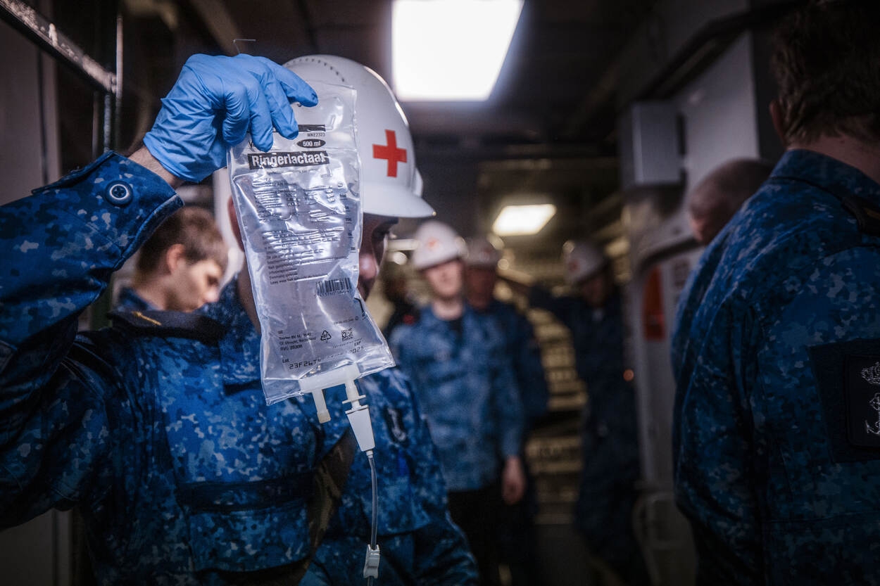 Een militair met een blauw marinepak en witte helm met daarop een rood kruis houdt een infuus in zijn handen.