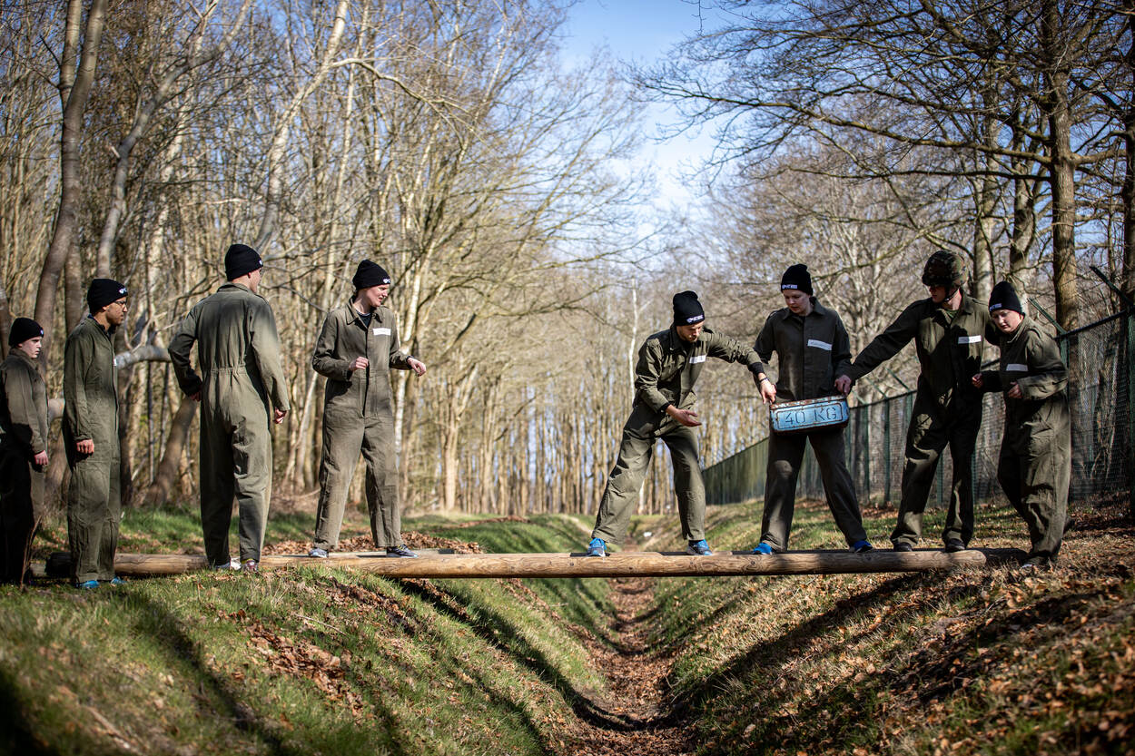 Een groep mensen, gekleed in groene overall, balanceert op een balk, die een sloot overbrugt. Samen proberen ze een munitiekist van 40 kilo van de ene naar de andere kant te tillen.