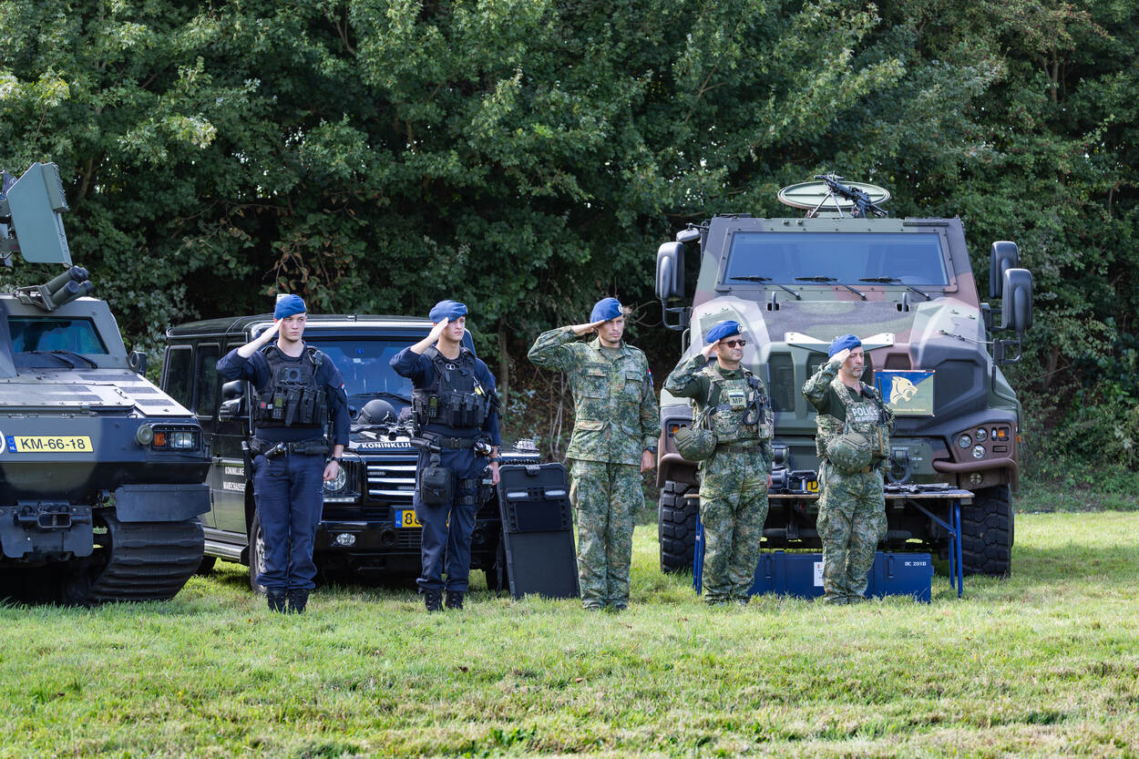 5 militairen salueren, achter ze staan voertuigen opgesteld.