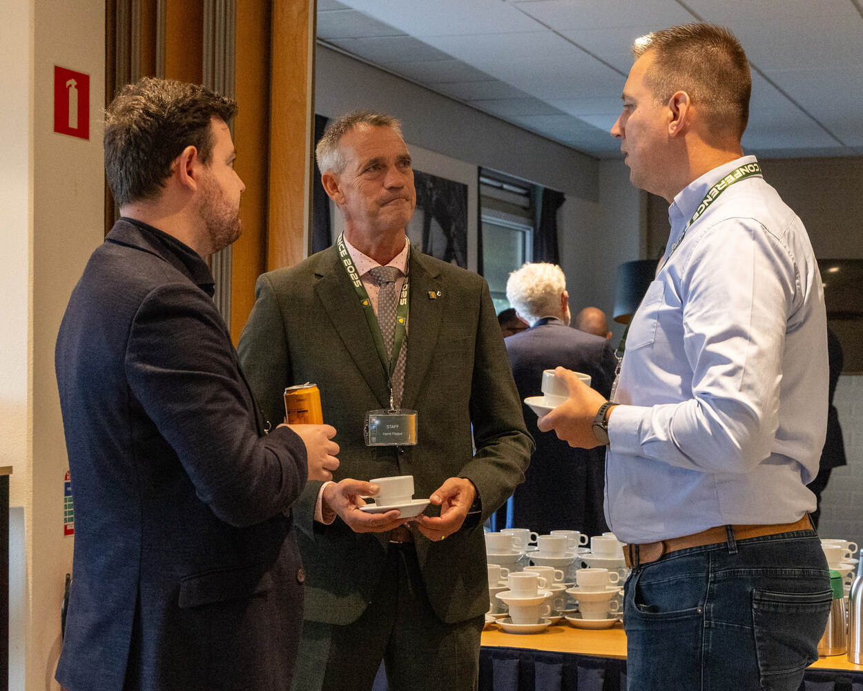 Drie heren met drinken in handen staan te kletsen voor een koffietafel.