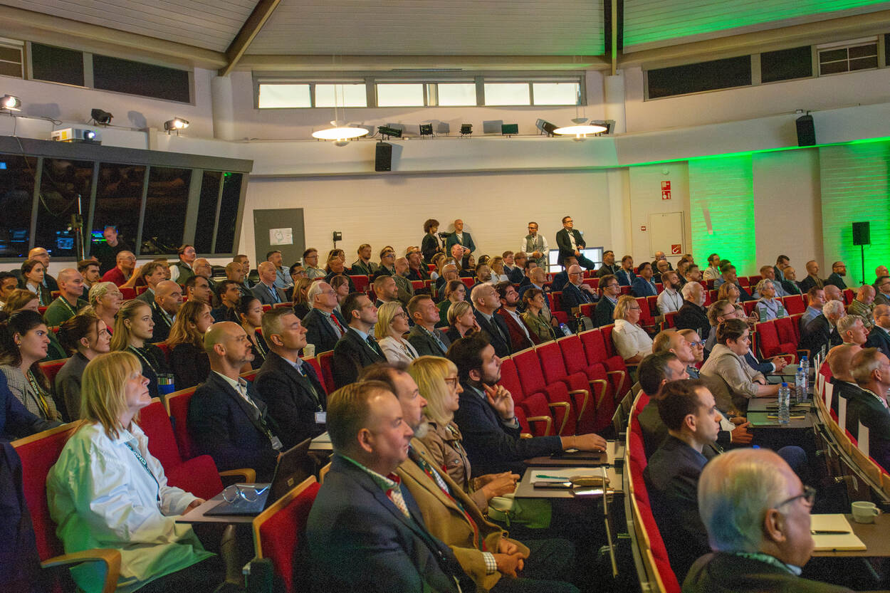 Meerdere mensen luisteren aandachtig in verlichte congreszaal.