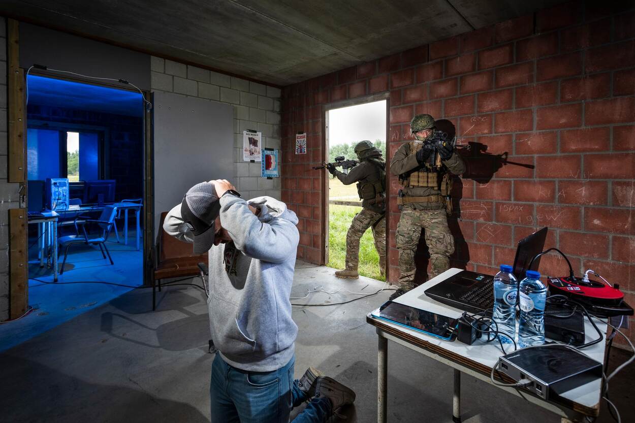 Twee militairen stappen met geweren een ruimte binnen waar een verdachte geknield op de grond zit. Om hem heen computers en andere IT-middelen.