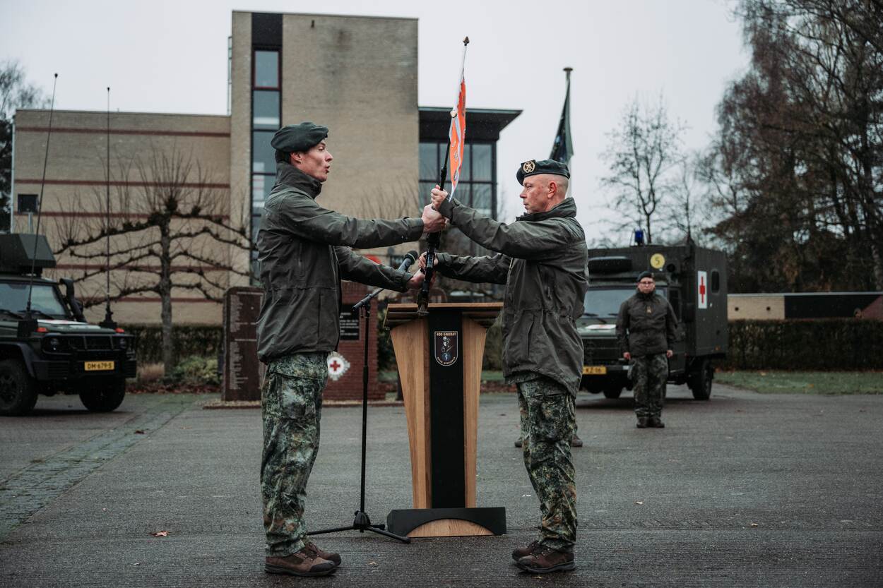 Twee militairen staan tegenover elkaar met een militaire vlag in het midden. Op de achtergrond staan een militair en twee militaire voertuigen.