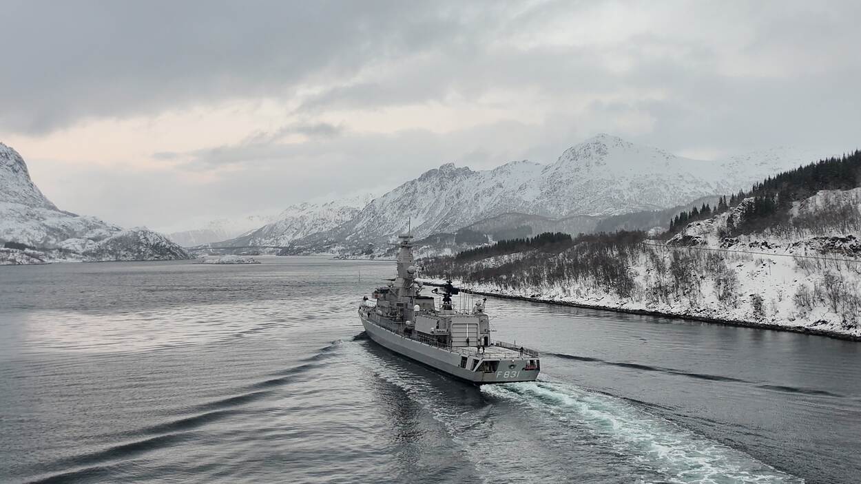 Een marinefregat vaart in een fjord waar sneeuw ligt.