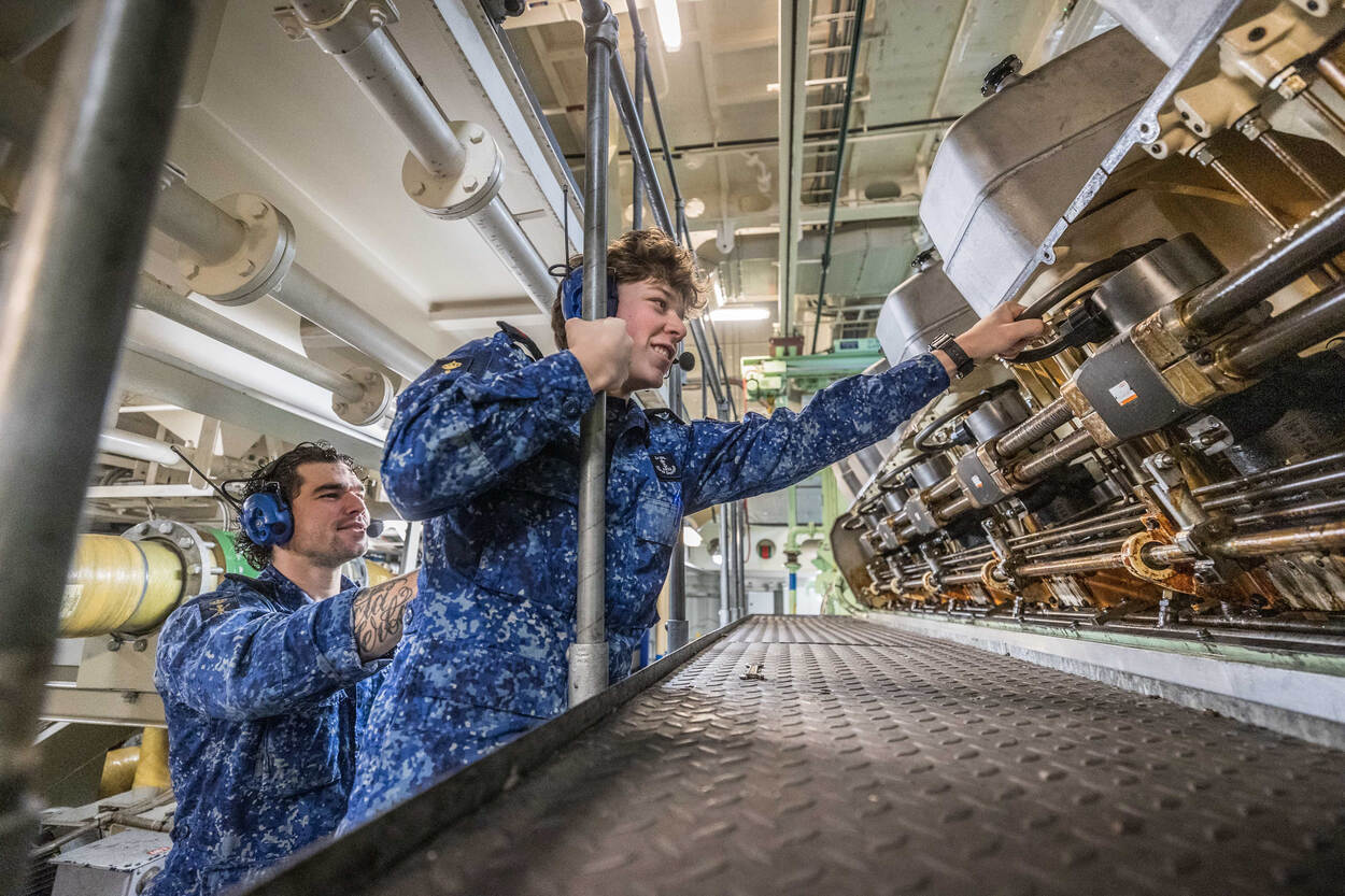 Een lachende matroos werkt in de technische ruimte van het schip, terwijl een collega hem ondersteunt.