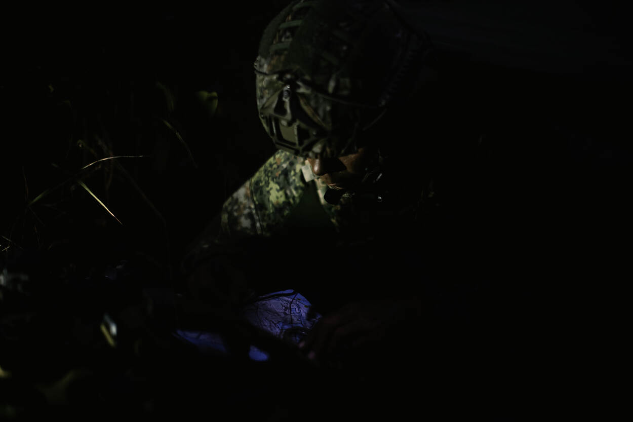 Een marinier kijkt naar apparatuur, dat hij in zijn handen vasthoudt. Hij draagt een  helm en ligt onder een zeiltje, in de ochtendgloren.