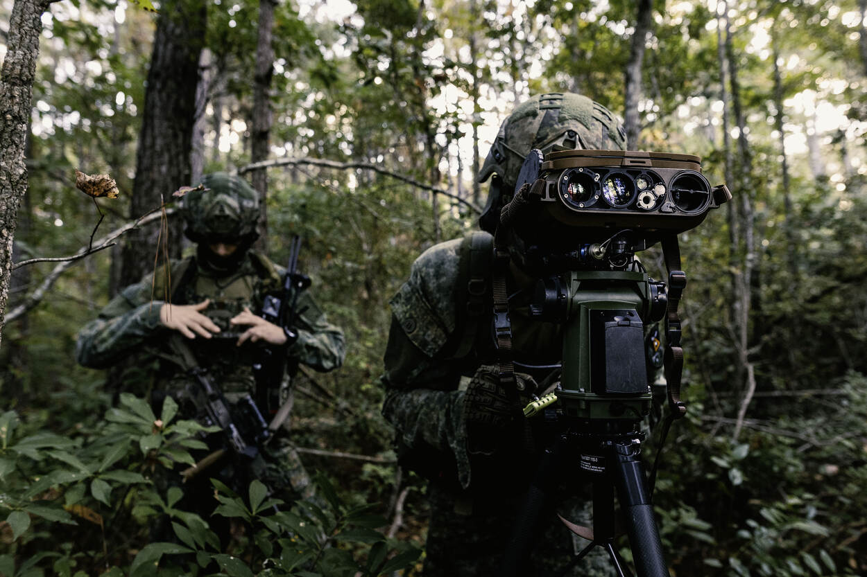 Een marinier tuurt door een gyroscoop, staand in het bos. Zijn collega achter hem kijkt naar een scherm dat voor zijn borst hangt.
