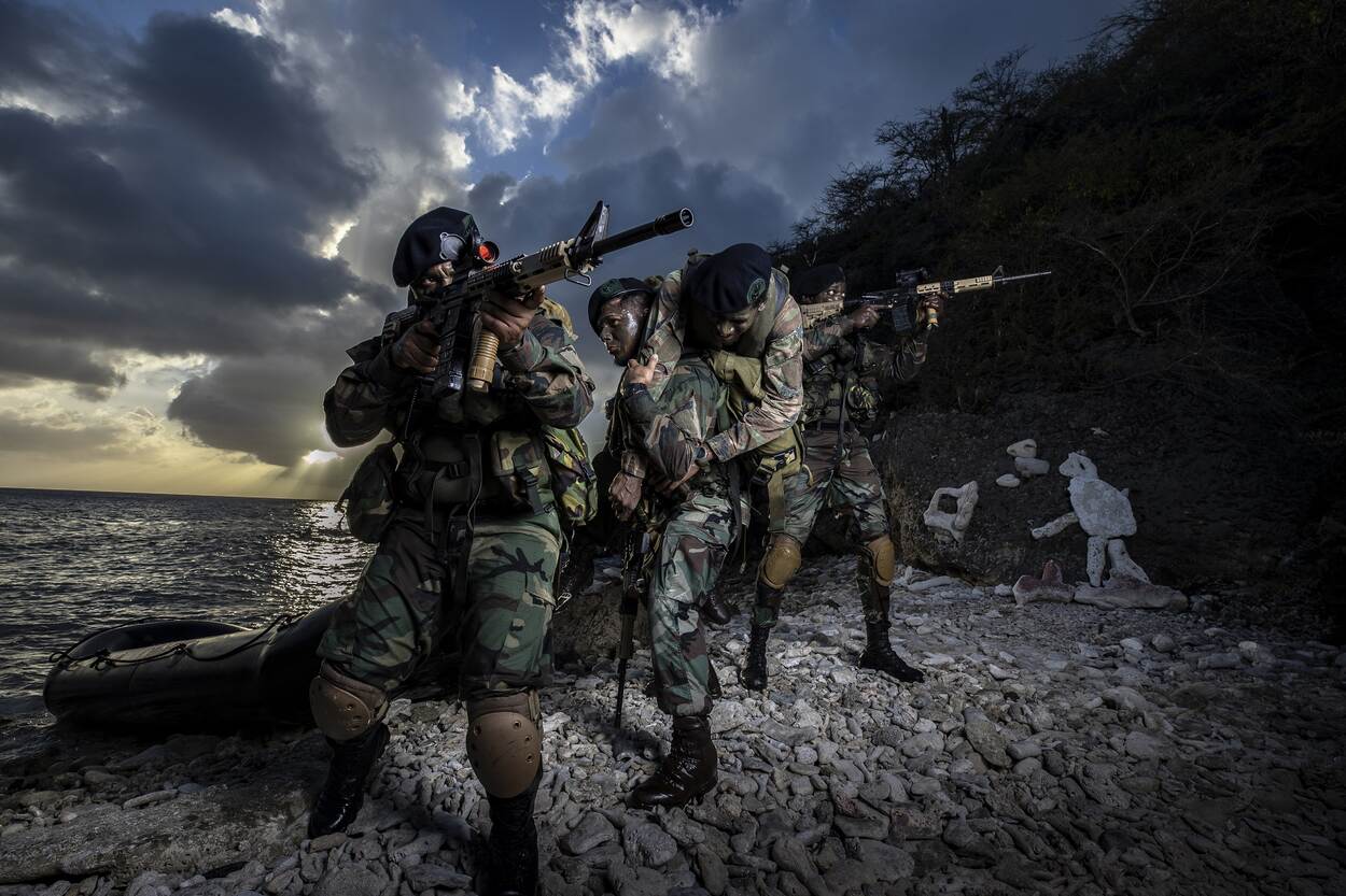 Caribische Militairen die met het geweer in de aanslag nabij een motorboot een rondom-verdediging uitvoeren om een gewonde collega van het strand te redden.
