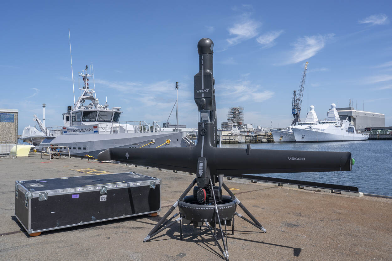 De zwartgekleurde V-Bat drone. Deze staat rechtop. Op de achtergrond liggen marineschepen in de haven.