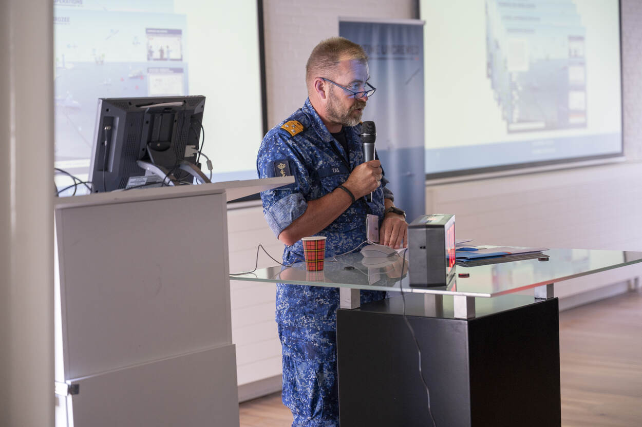 C-ZSK vice-admiraal René Tas spreekt de zaal toe door een microfoon. Achter hem hangen 2 schermen.