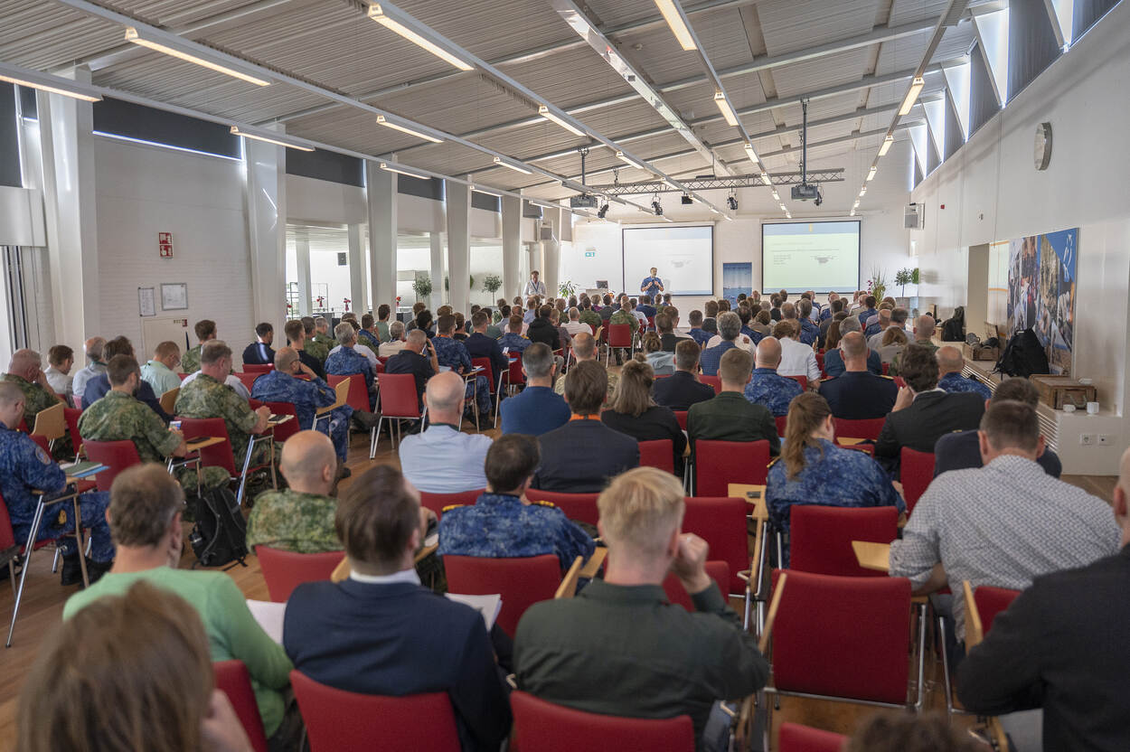 Een groot aantal aanwezigen luistert op rode stoelen naar een van de sprekers.