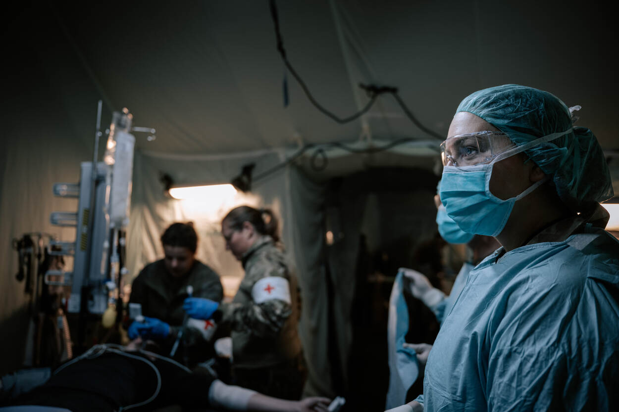 Binnenkant van een militair hospitaal te velde. Op de voorgrond staat een militair met een operatie-outfit aan. Op de achtergrond zien we een gewonde militair op een bed liggen, die wordt verzorgd door twee genezerikken.