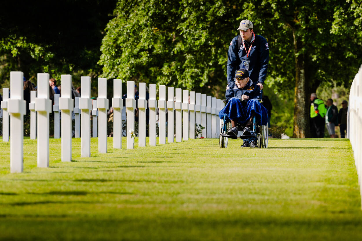 Een veteraan wordt in een rolstoel vooruitgeduwd tussen de rijen met witte grafstenen.