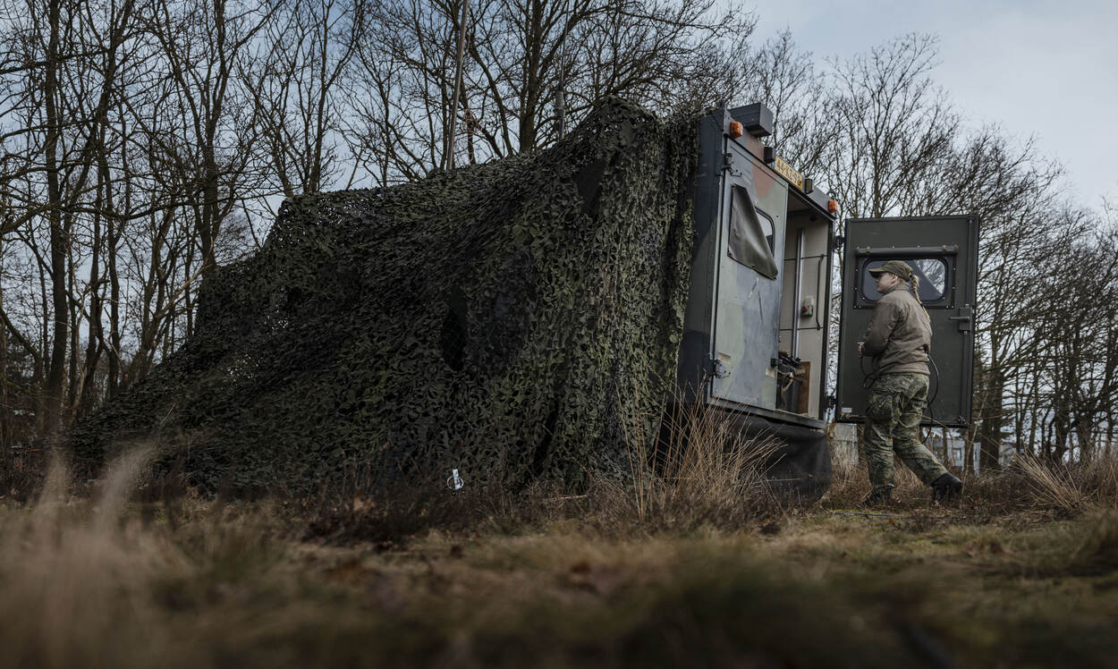 Een militair staat op de hei voor de ingang van een gecamoufleerd militair voertuig.