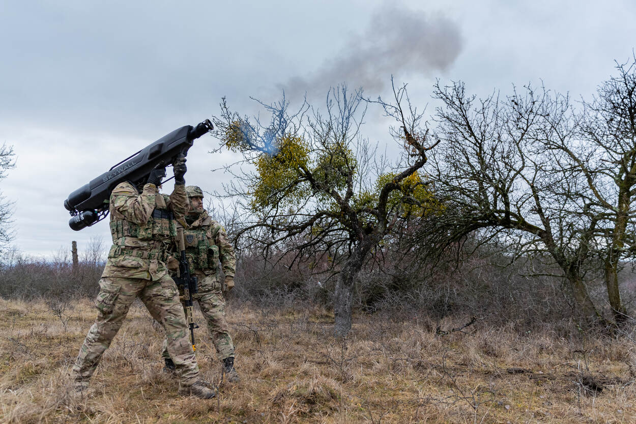 Op de ene foto vliegt een drone door de lucht. Door de beweging, is de achtergrond vaag. Op de andere foto zijn twee militairen te zien, met een groot langwerpig apparaat in handen. De rookpluimen komen uit de loop van de zwarte dronevanger.
