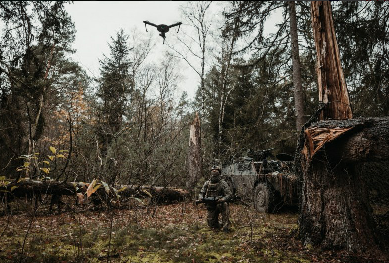 Een militair kijkt omhoog. Boven hem vliegt een drone, die hij met het console in de handen bestuurt. Om hem heen staan voertuigen en bomen.