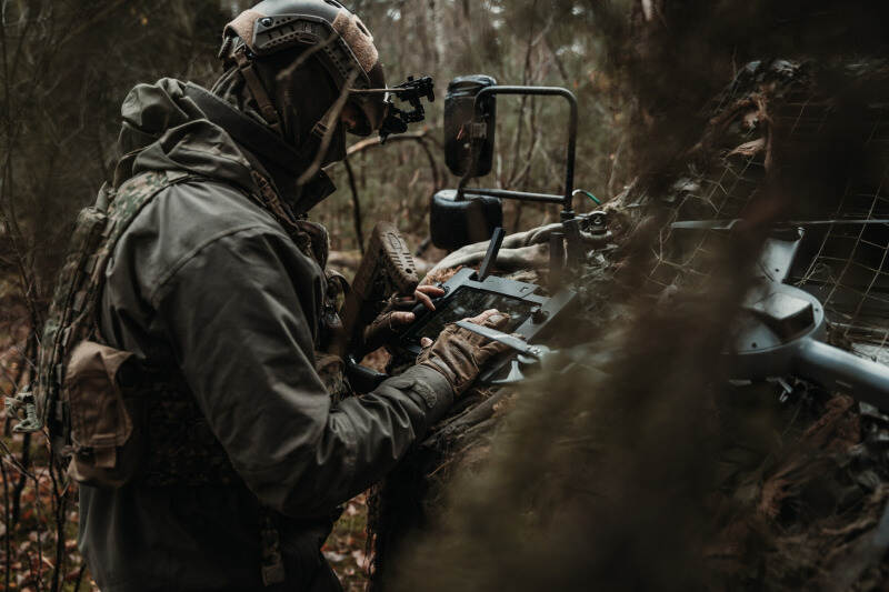 Een onherkenbare militair buigt over een droneconsole, staand vlak voor een voertuig aan de bosrand.