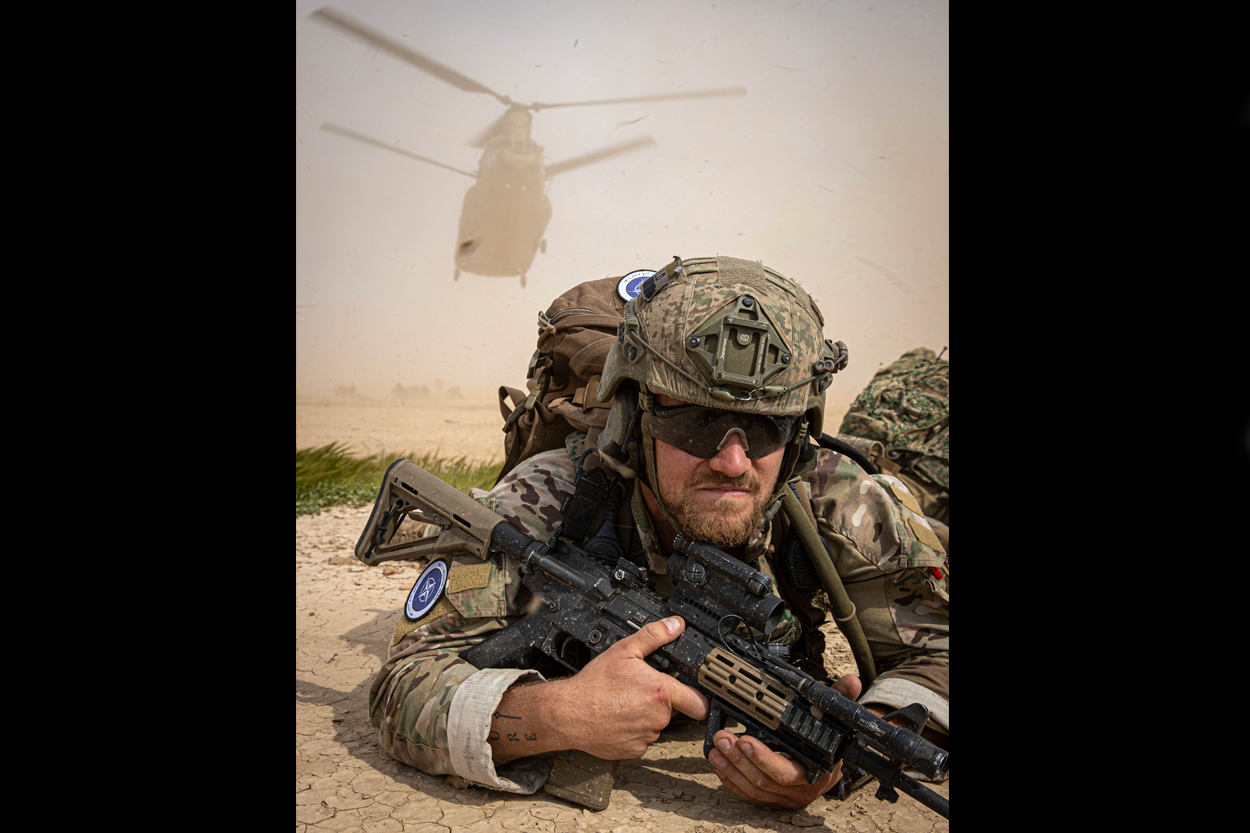 Een Nederlandse militair van 11 Luchtmobiele Brigade ligt op de grond, terwijl achter hem een Chinook-helikopter landt.
