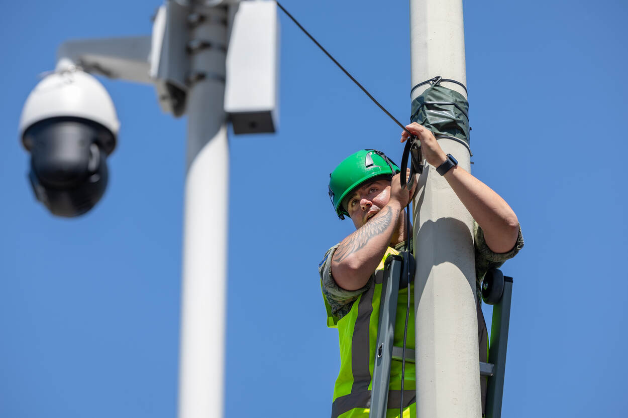 Militair in hesje en helm op legt vanaf ladder kabel aan.