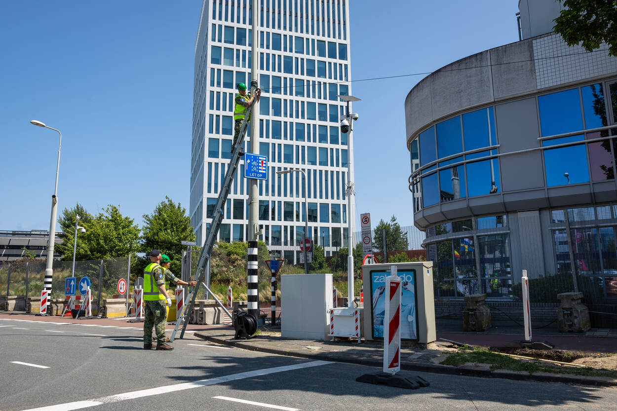 2 militairen in hesje en met helm op ondersteunen een ladder voor collega die kabel aanlegt.