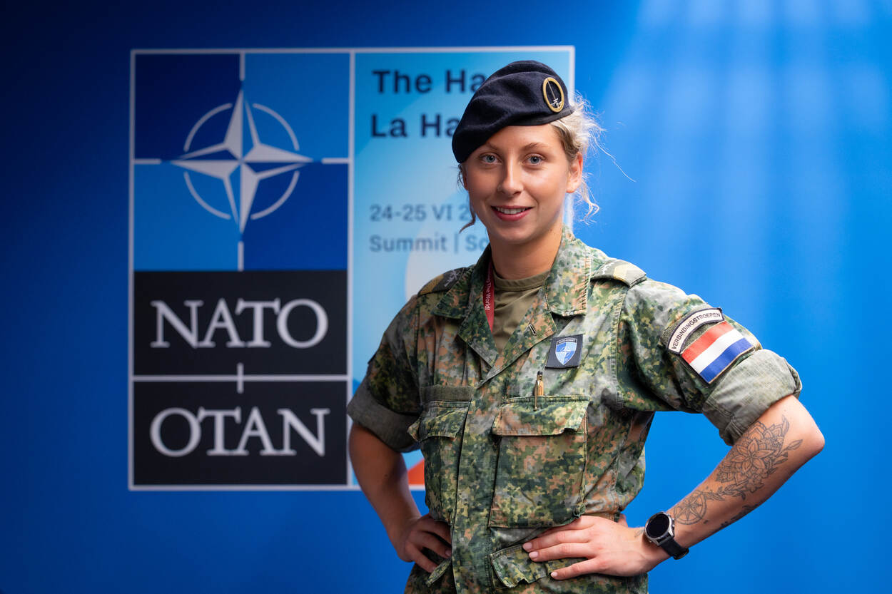 Vrouwelijke korporaal met baret poseert voor logo NAVO-top.