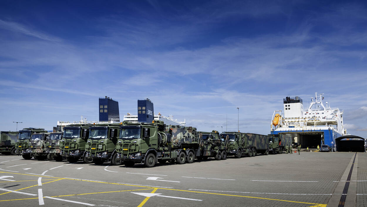 Militaire trucks staan op de kade, met op de achtergrond een transportschip.