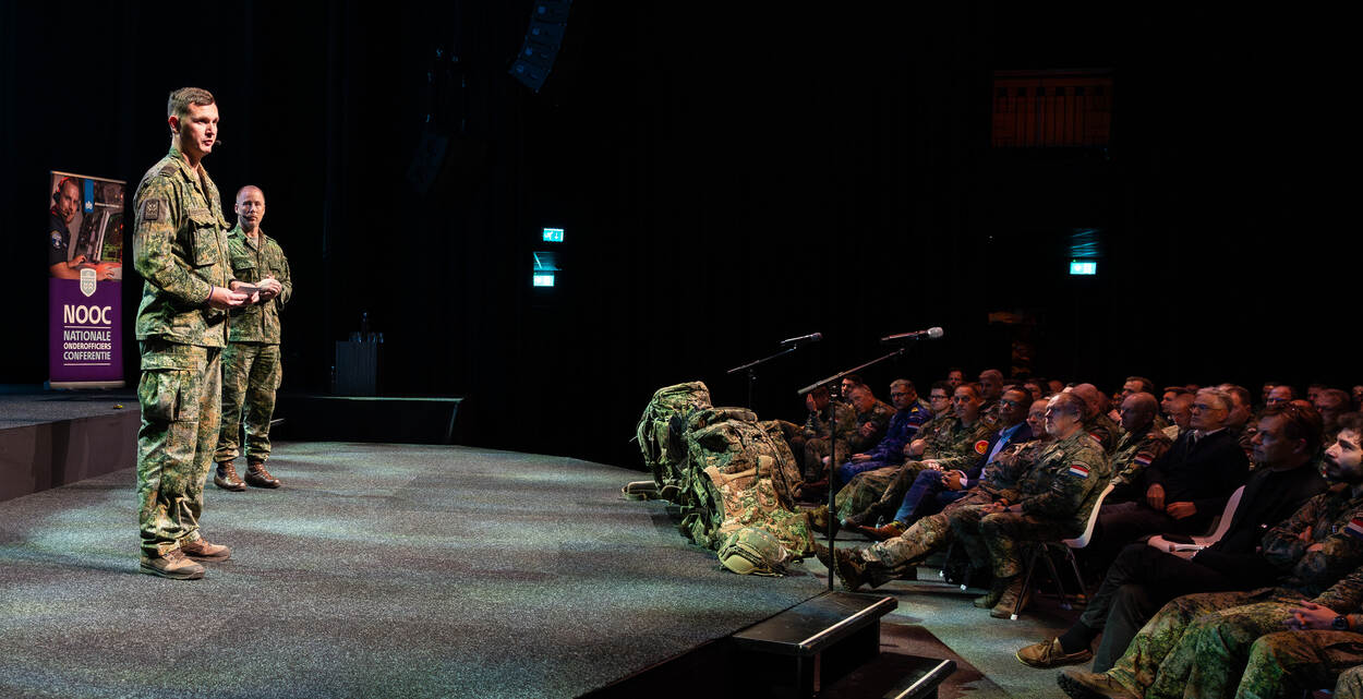 Een sergeant-majoor van de luchtmacht spreekt op het podium de zaal toe.