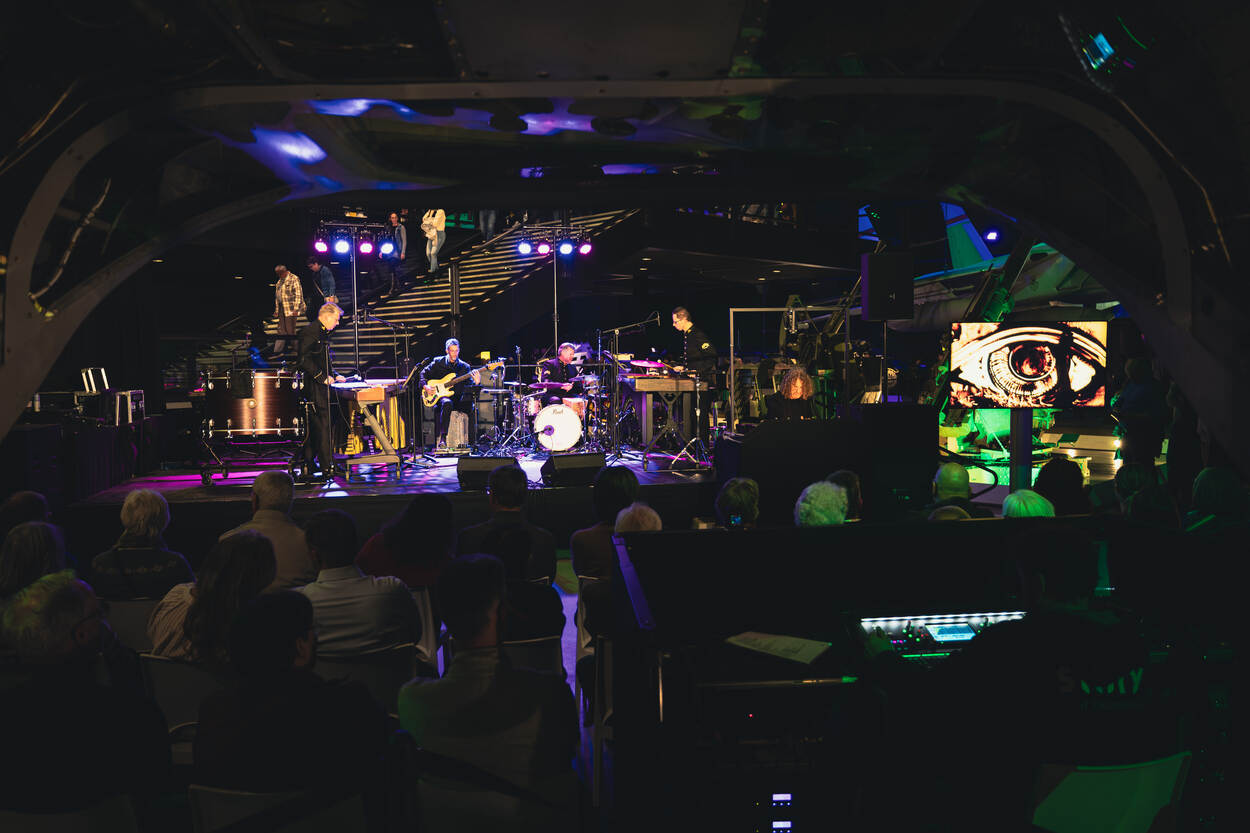 Slagwerkers van het Orkest Koninklijke Luchtmacht met rechts daarnaast de zandtovenaar die van zand een oog maakt.