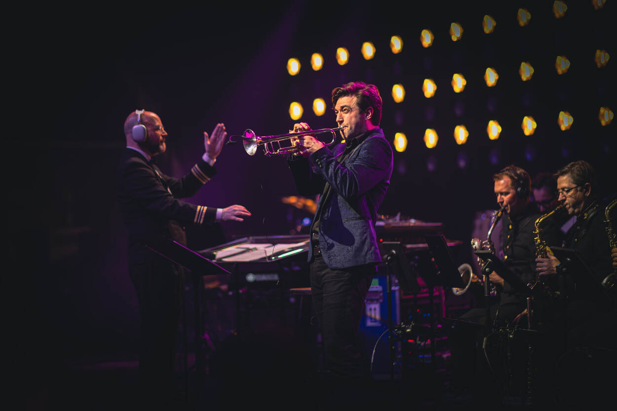 Een burger solotrompettist tijdens een optreden met het Orkest Koninklijke Luchtmacht met op de achtergrond links de dirigent en rechts twee blazers van het orkest.
