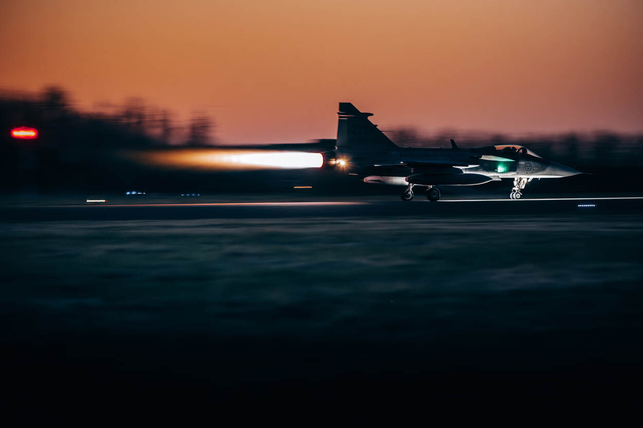 Een Zweedse Gripen vertrekt met naverbrander vanaf Leeuwarden tegen een rode avondlucht.