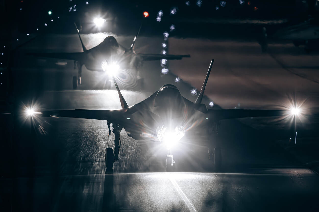 Drie F-35’s taxiën in het donker. Felle lampen schijnen vanaf hun landingsgestel op de taxibaan.