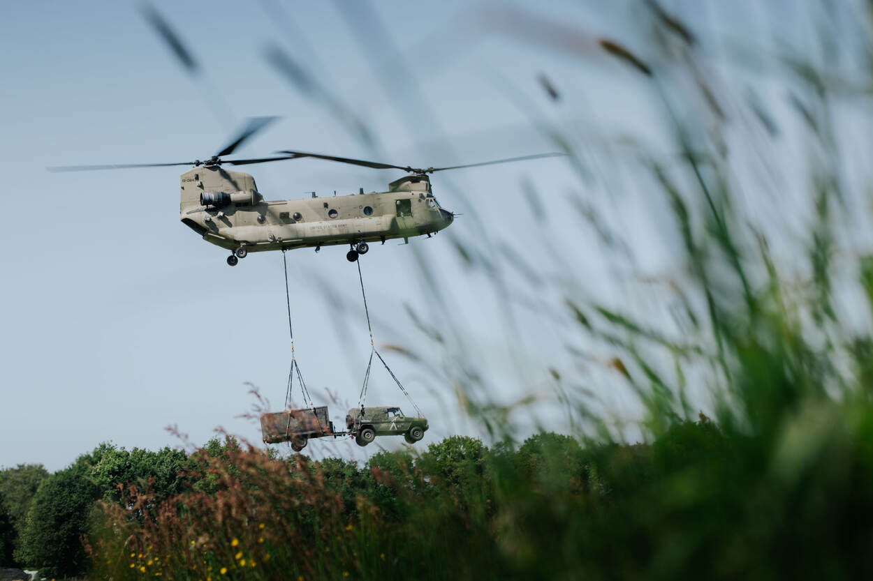 Een vliegende Chinook met daaronder hangend een jeep met aanhanger.