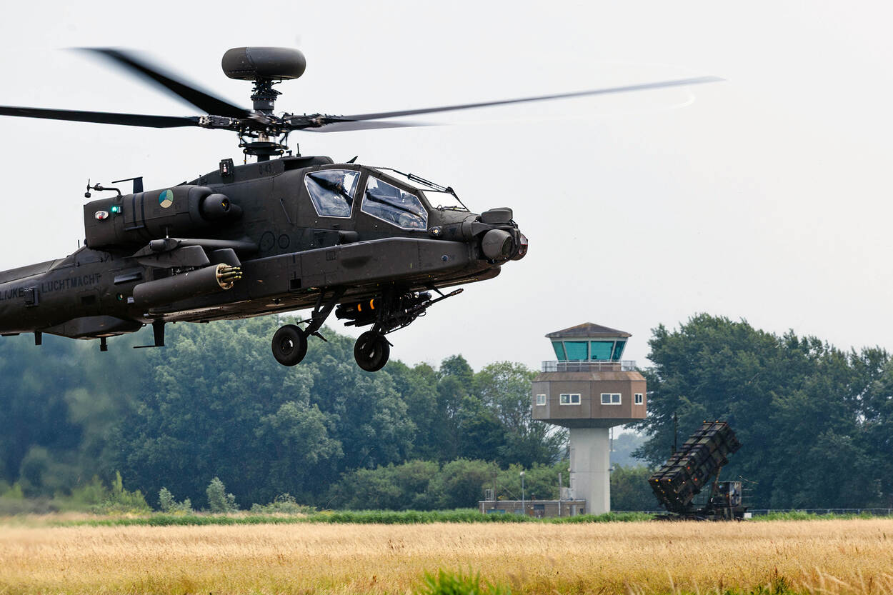 Gevechtshelikopter vliegt vlak boven de grond met op de achtergrond een verkeerstoren en Patriot-luchtverdedigingssyteem.