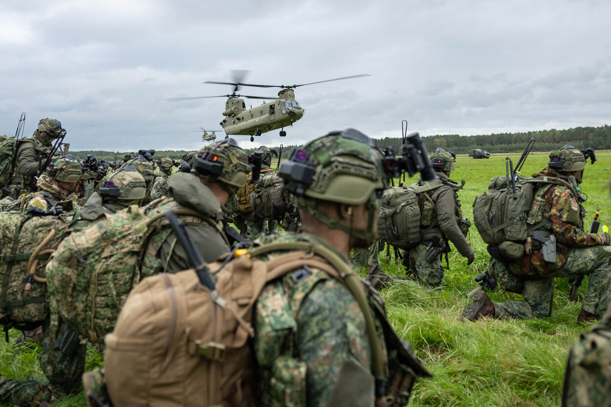 Twee Chinook vliegen boven een groep militairen die geknield op de grond zitten.