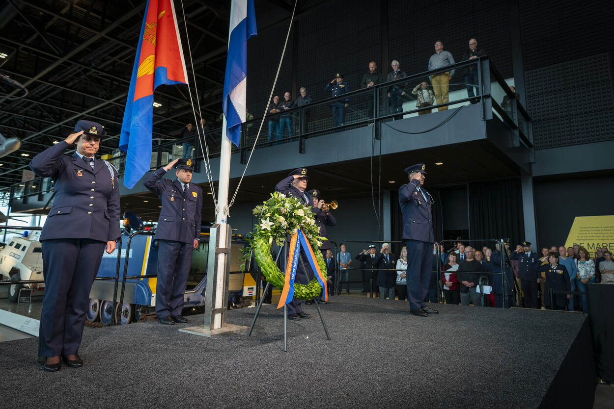 Een krans met daarachter de Nederlandse vlag en luchtmachtvlag halfstok, en luchtmachtmilitairen in de houding.