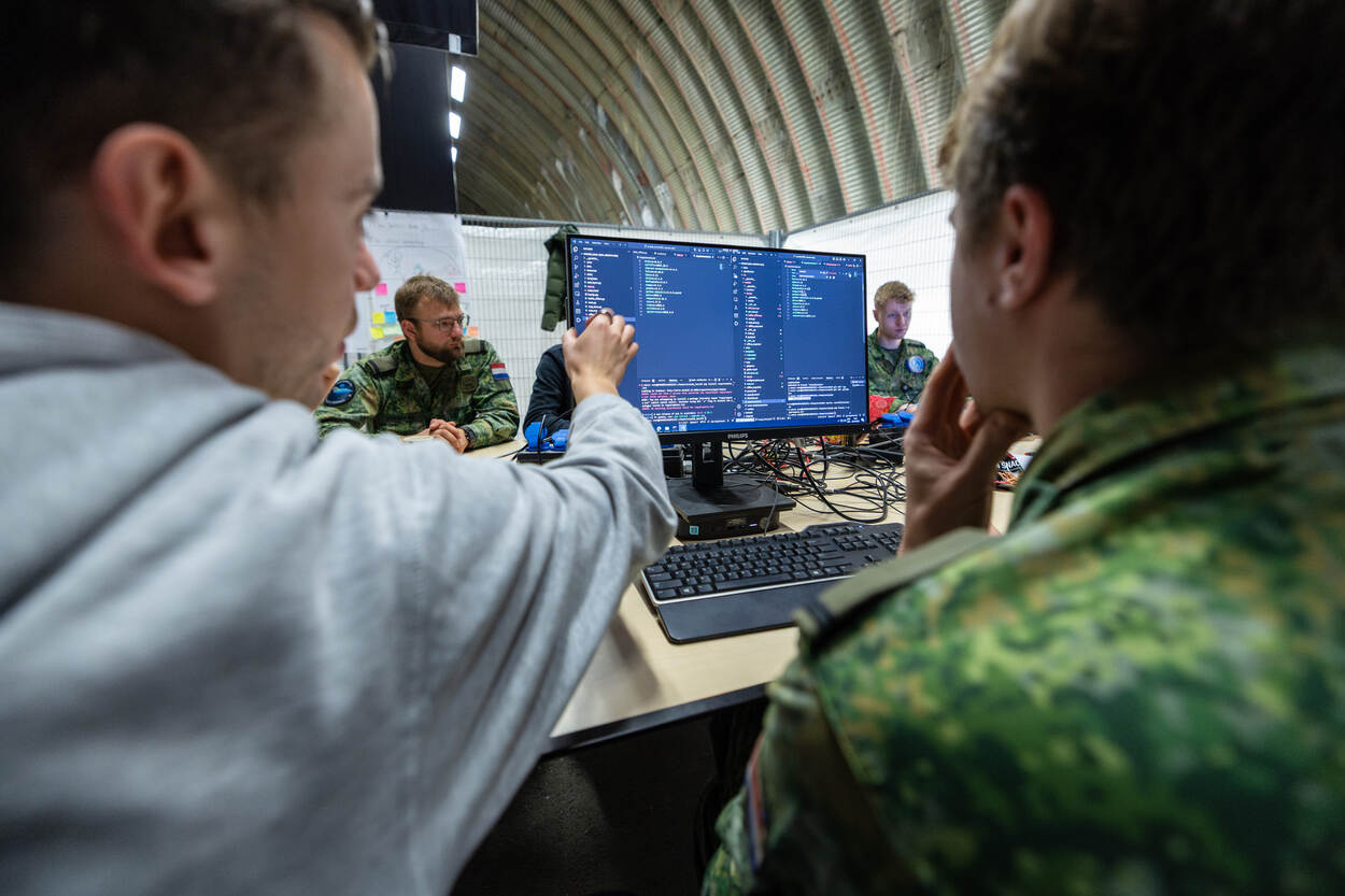 Twee personen, een militair en burger, zitten naast elkaar en kijken naar een computerscherm. Daarop is een softwarecode te zien.