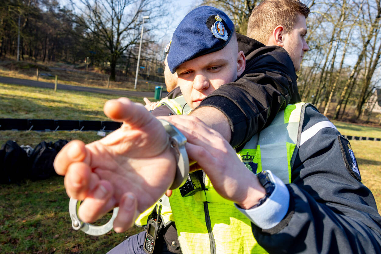 Twee marechaussees staan met hun rug naar elkaar toe. Daartussen staat een tegenspeler, die in de boeien wordt geslagen.