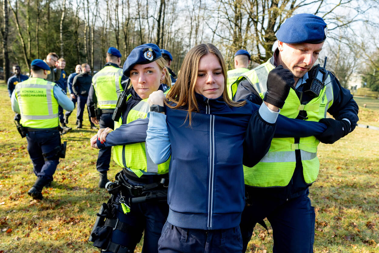 Twee marechaussees verplaatsen een tegenspeelster in een blauw pak door ieder een van hun armen onder haar oksel te plaatsen, en met hun vrije hand haar polsen vast te houden.