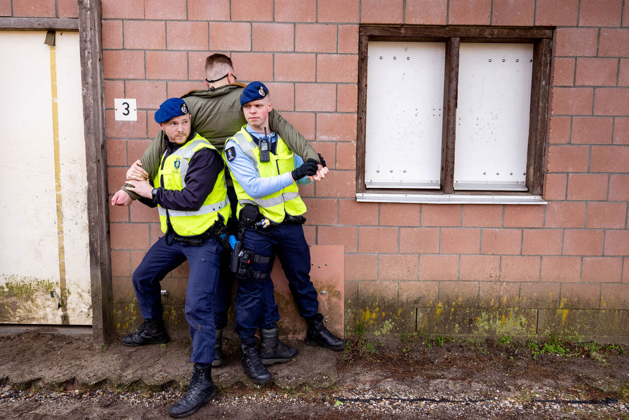 Twee marechaussees drukken een tegenspeler met zijn buik tegen een muur. Omdat ze zelf omgekeerd staan, kunnen ze de omgeving in de gaten houden.