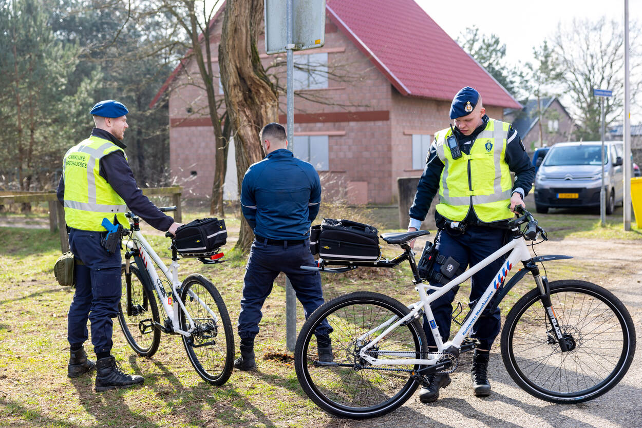 Een tegenspeler staat te plassen tegen een verkeersbord, daaromheen twee marechaussees met fietsen.