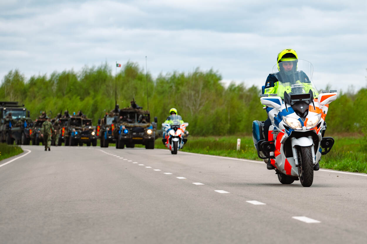 Twee motorrijders rijden voor een militaire colonne uit.