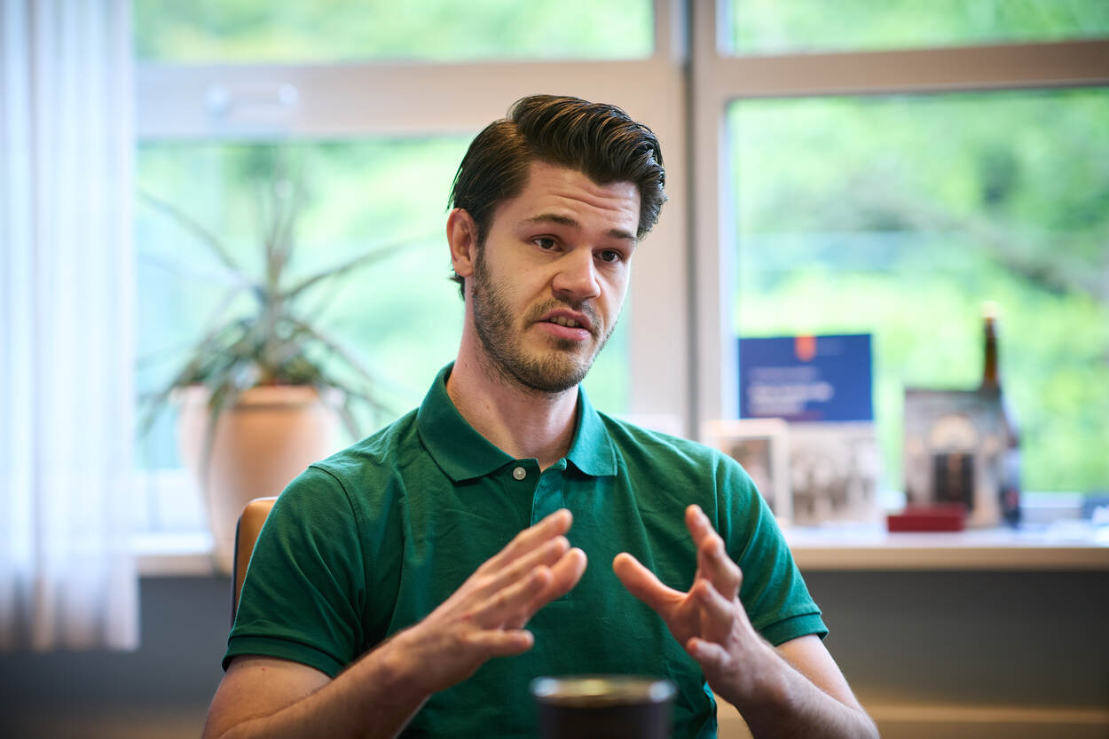 Een burger in groen shirt doceert voor een raam met op de achtergrond een plant en fotolijsten.