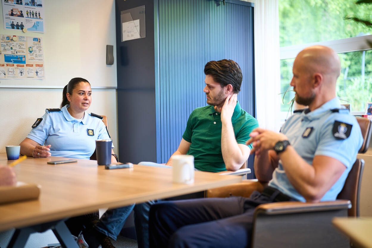 Een majoor van de KMar neemt aan tafel het woord, terwijl een collega en een burger luisteren.