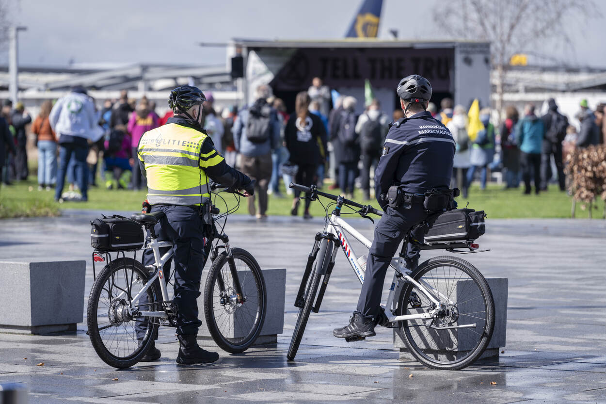 Een politieagent en marechaussee, zittend op hun fietsen. Beiden hebben ze hun benen op de grond. Op de achtergrond is een mensenmenigte te zien.