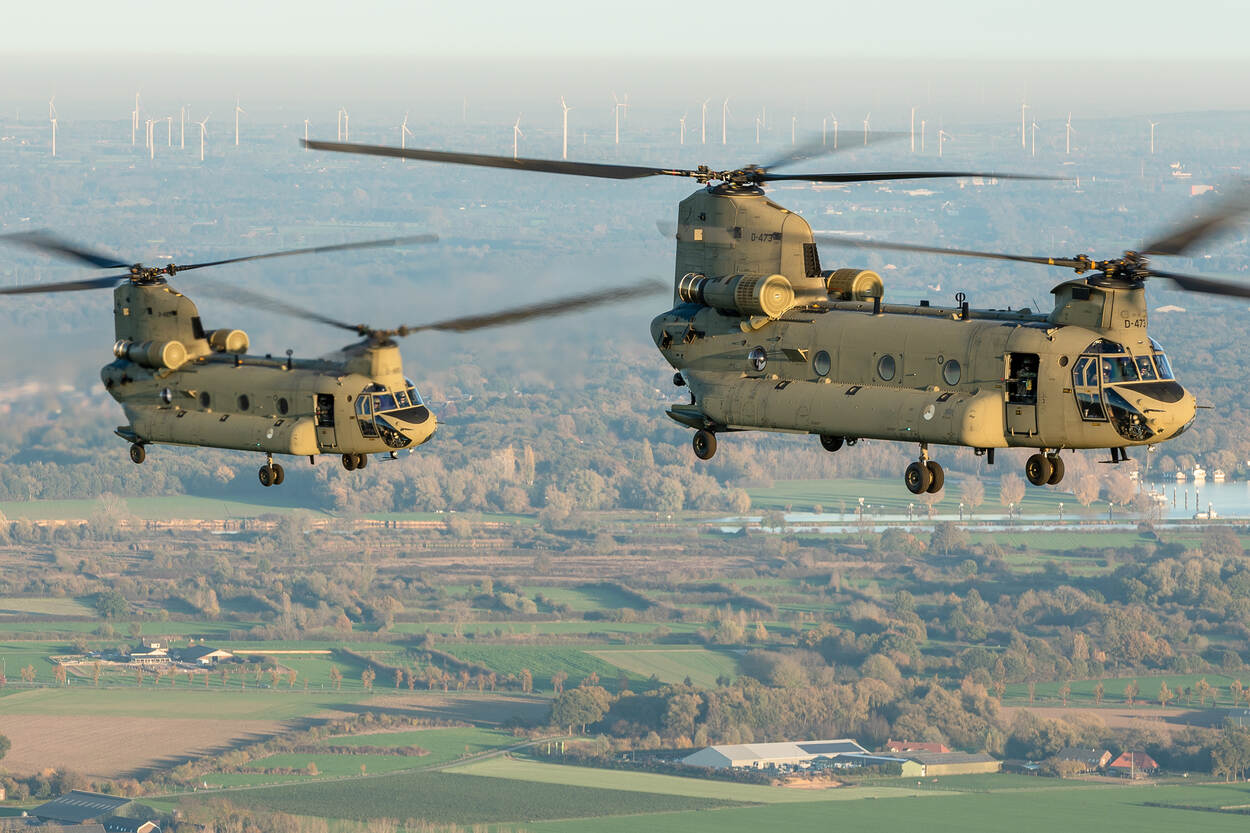 Twee Chinook CH-47F transporthelikopters boven een Nederlands landschap met op de achtergrond een windmolenpark.