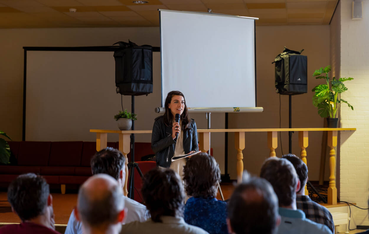 Een vrouw met microfoon spreekt een zaal toe met op achtergrond 2 videoschermen.